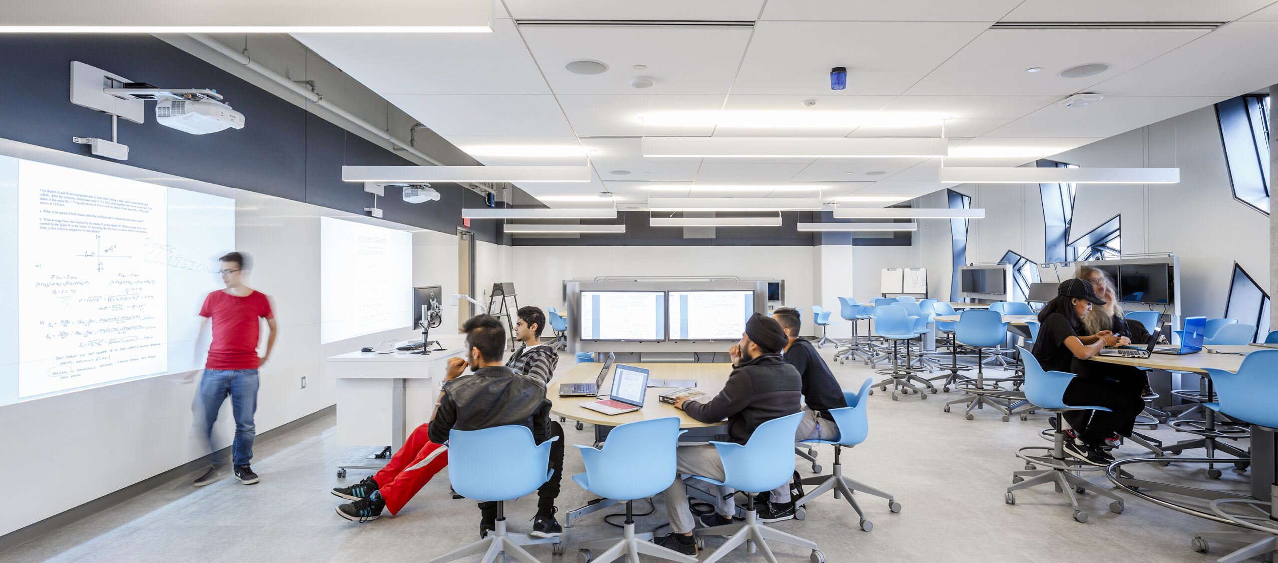 wide view of the Lassonde classroom interior space, diverse young people around a table listening to speaker in front of projector screen