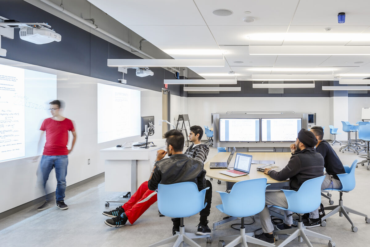 Lassonde classroom interior space, diverse young people around a table listening to speaker in front of projector screen