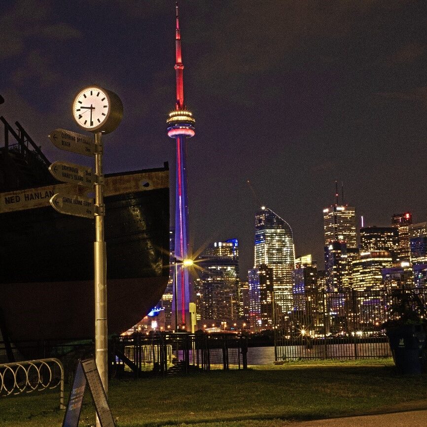 a rustic clock and a boat imposed against the Toronto skyline at night from Toronto Islands