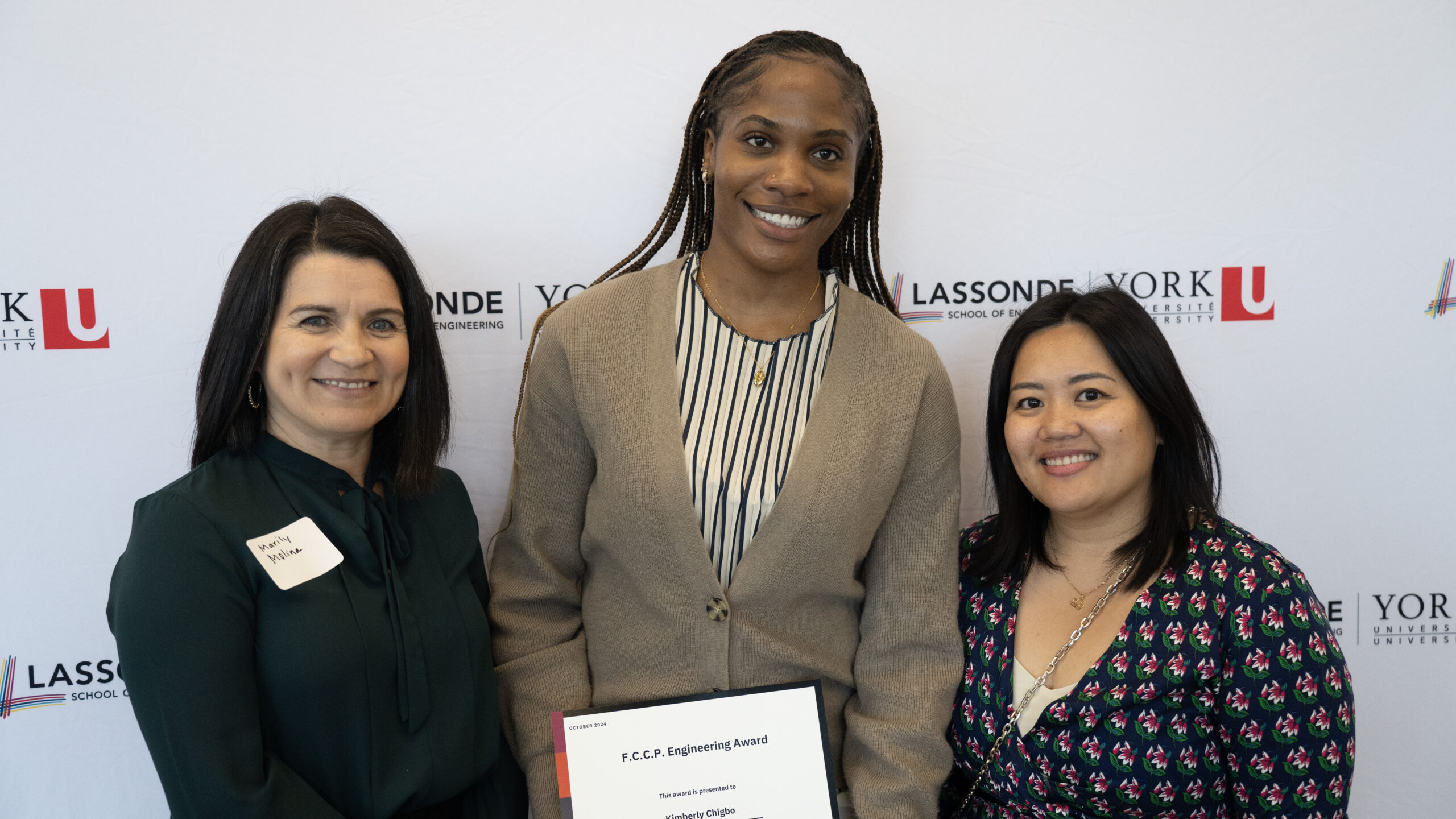 Three women posing for a photo in front of a Lassonde backdrop. The student in the middle is holding an award certificate.