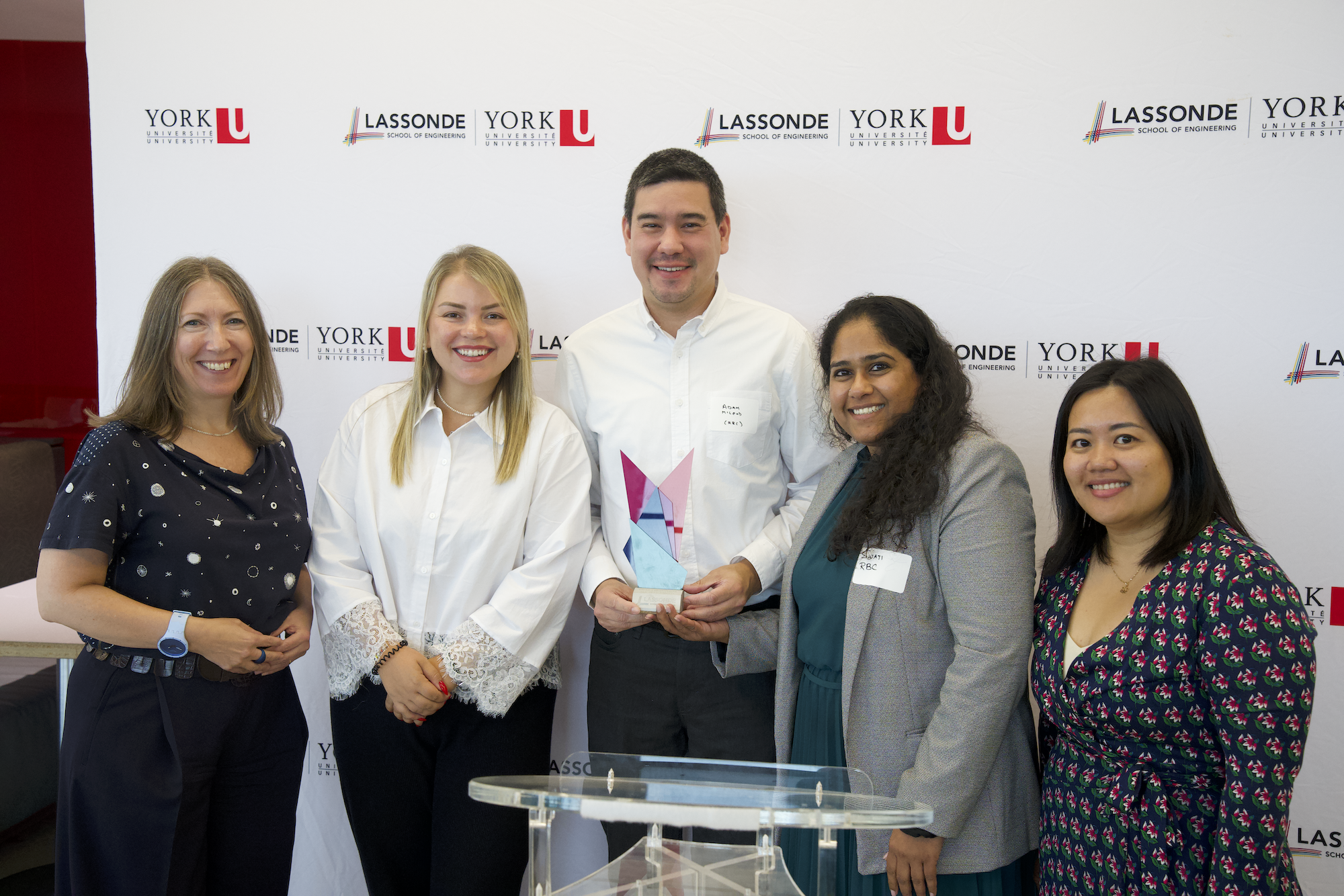 A group of people standing in front of a backdrop. The person in the centre is holding an award.