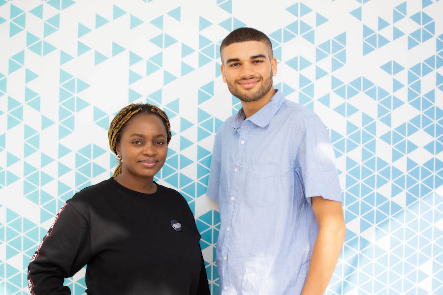A photo of a woman and a man smiling at the camera in front of a wall of blue triangles