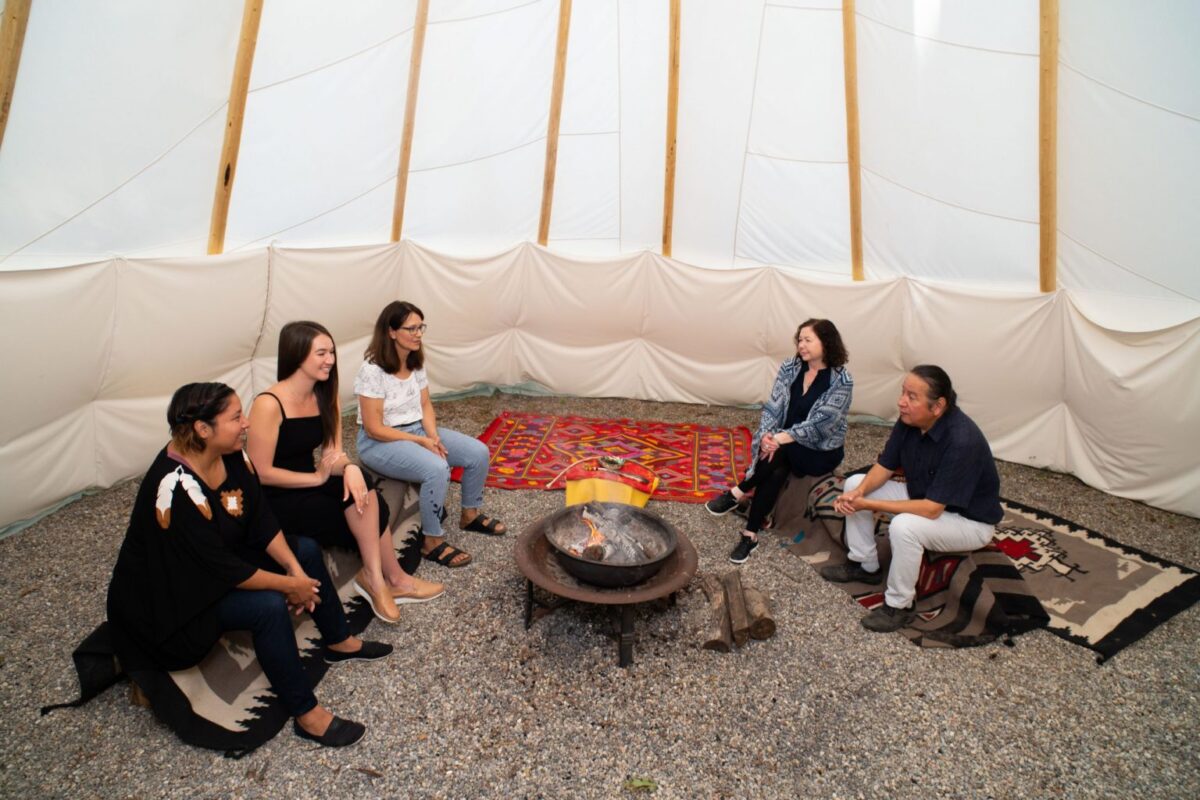 A group people sitting around a fire pit inside a traditional tipi hut