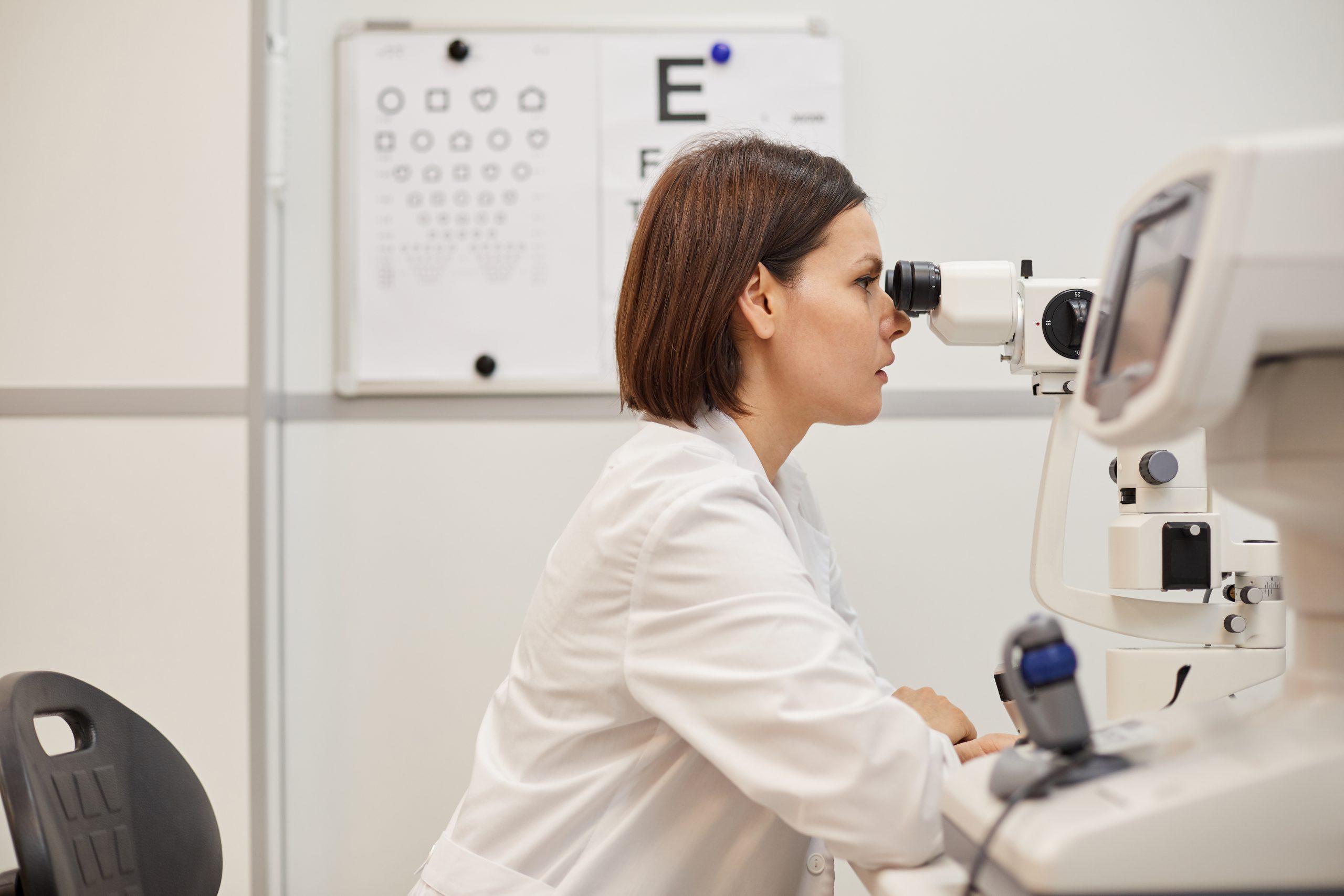 Side view portrait of young female ophthalmologist using refractometer machine during vision test in modern clinic, copy space